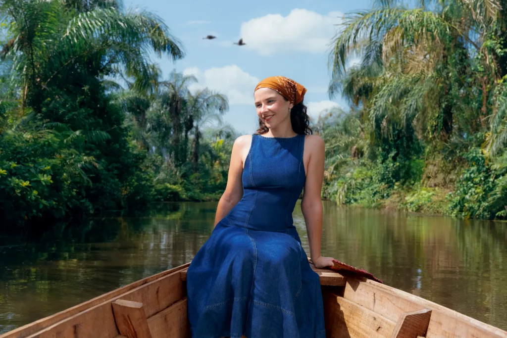 Uma mulher com um vestido azul e um lenço amarelo, sentada na proa de uma piroga, entre as ráfias e as palmeiras de ambas as margens