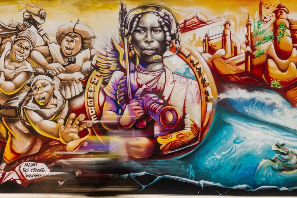 Détail d’une fresque murale aux couleurs vives : une figure féminine tient un appareil photo, entourée de visages et d’une vague