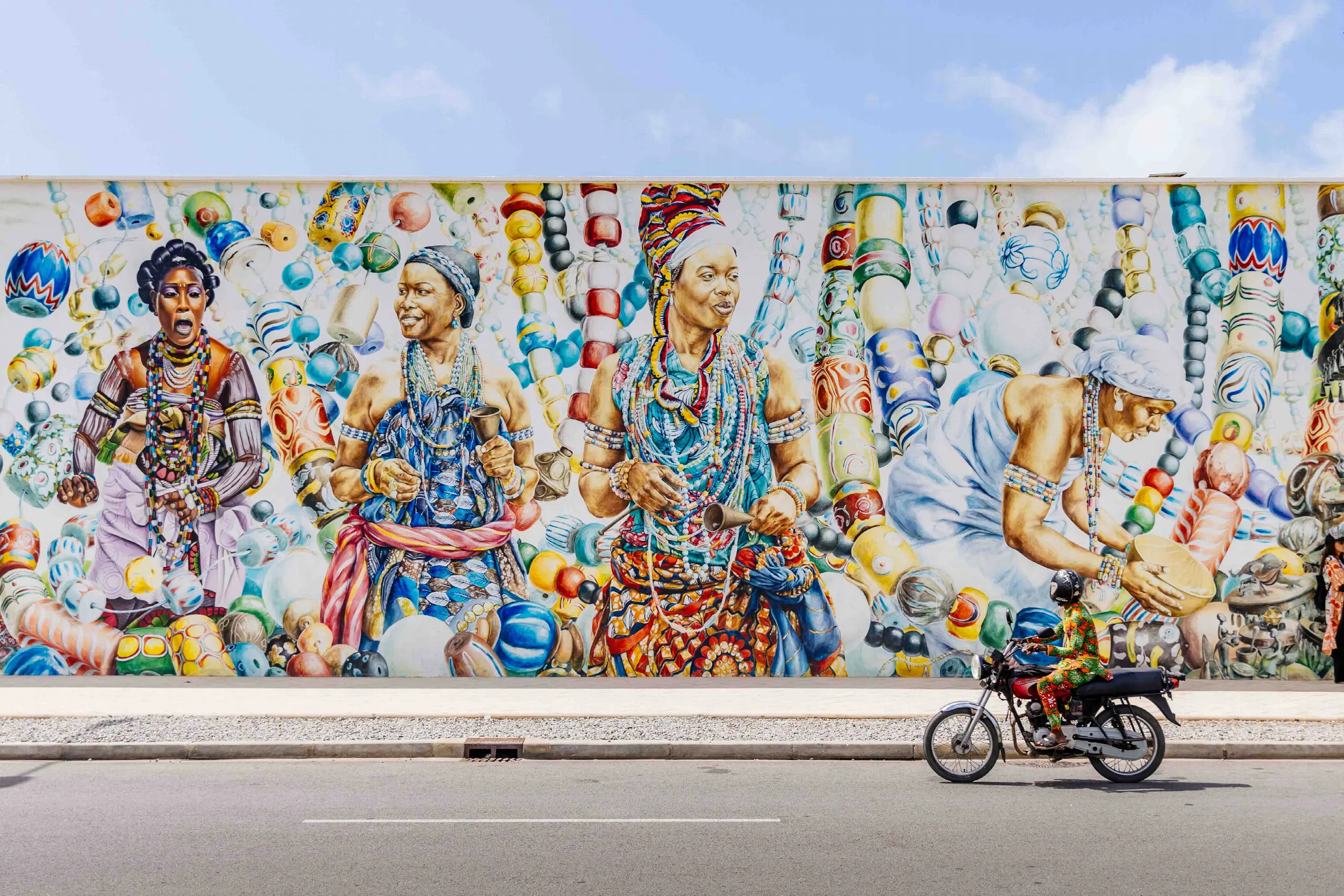 Immense fresque murale colorée représentant des femmes parées de perles et de pagnes, devant laquelle passe un motard