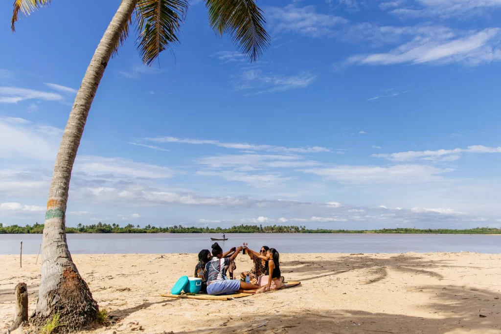Un groupe pique-nique sur une natte, sur le sable, sous un cocotier penché au-dessus de l’eau