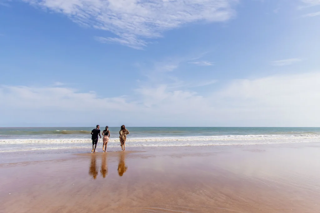 Trois promeneurs marchent au bord de l’eau sur une plage de sable humide, face à l’océan et sous un ciel bleu
