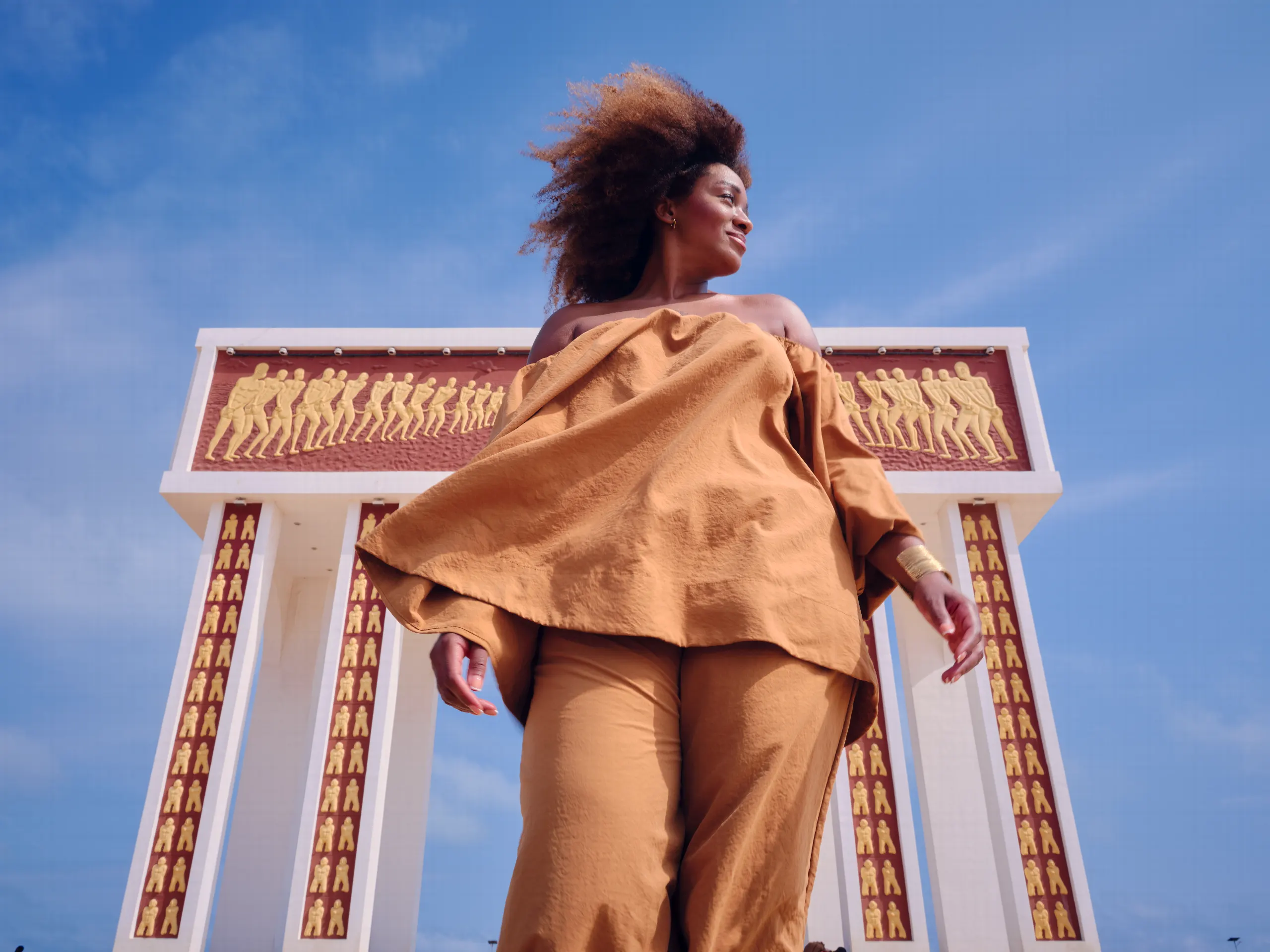 Une femme en tenue beige, les cheveux au vent, vue en contre-plongée devant un monument blanc orné de silhouettes en relief, sous un ciel bleu