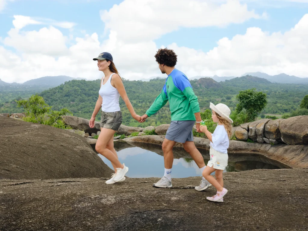 Une famille marche main dans la main sur une dalle de granit, près d’une vasque d’eau, avec des collines boisées derrière