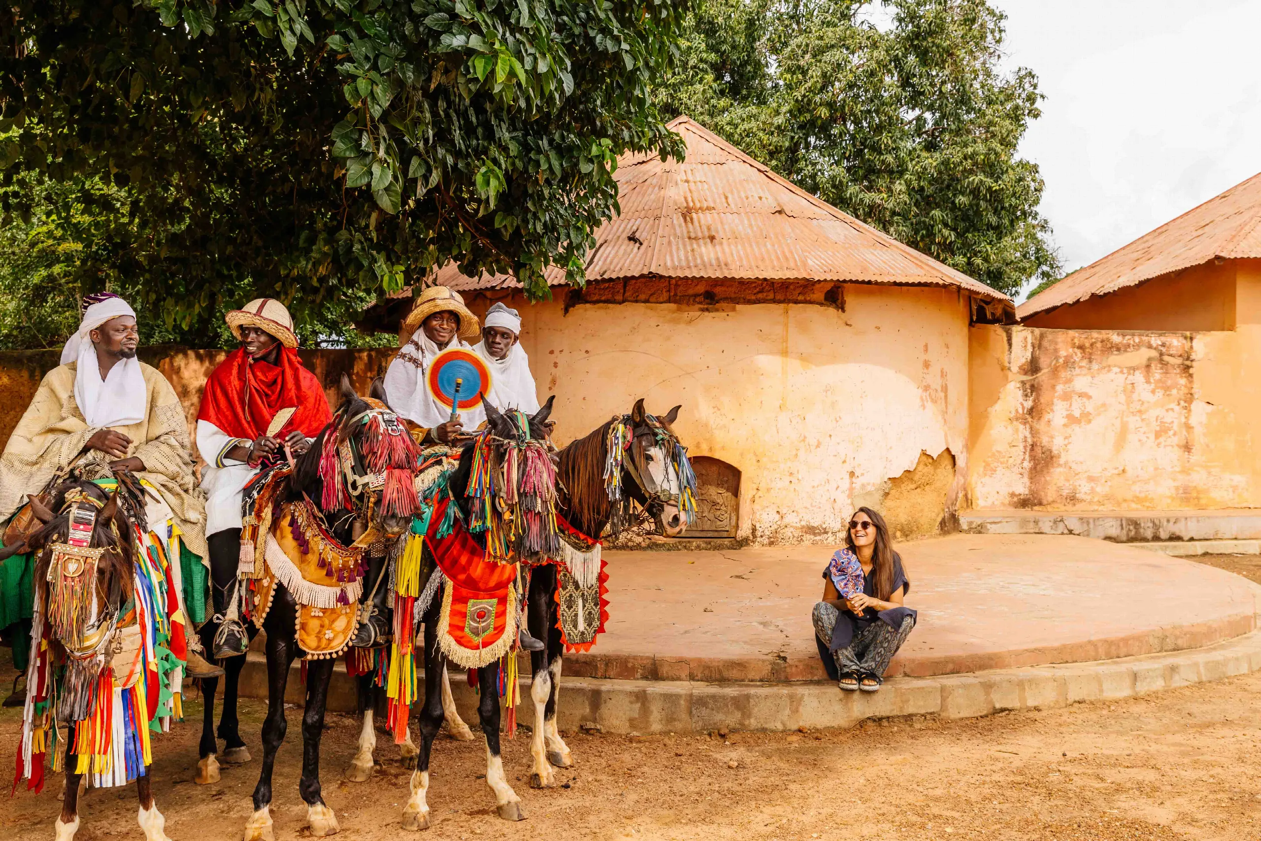 Des cavaliers en tenue d’apparat sur des chevaux ornés de franges colorées, devant un bâtiment de terre ocre, tandis qu’une visiteuse est assise sur un muret