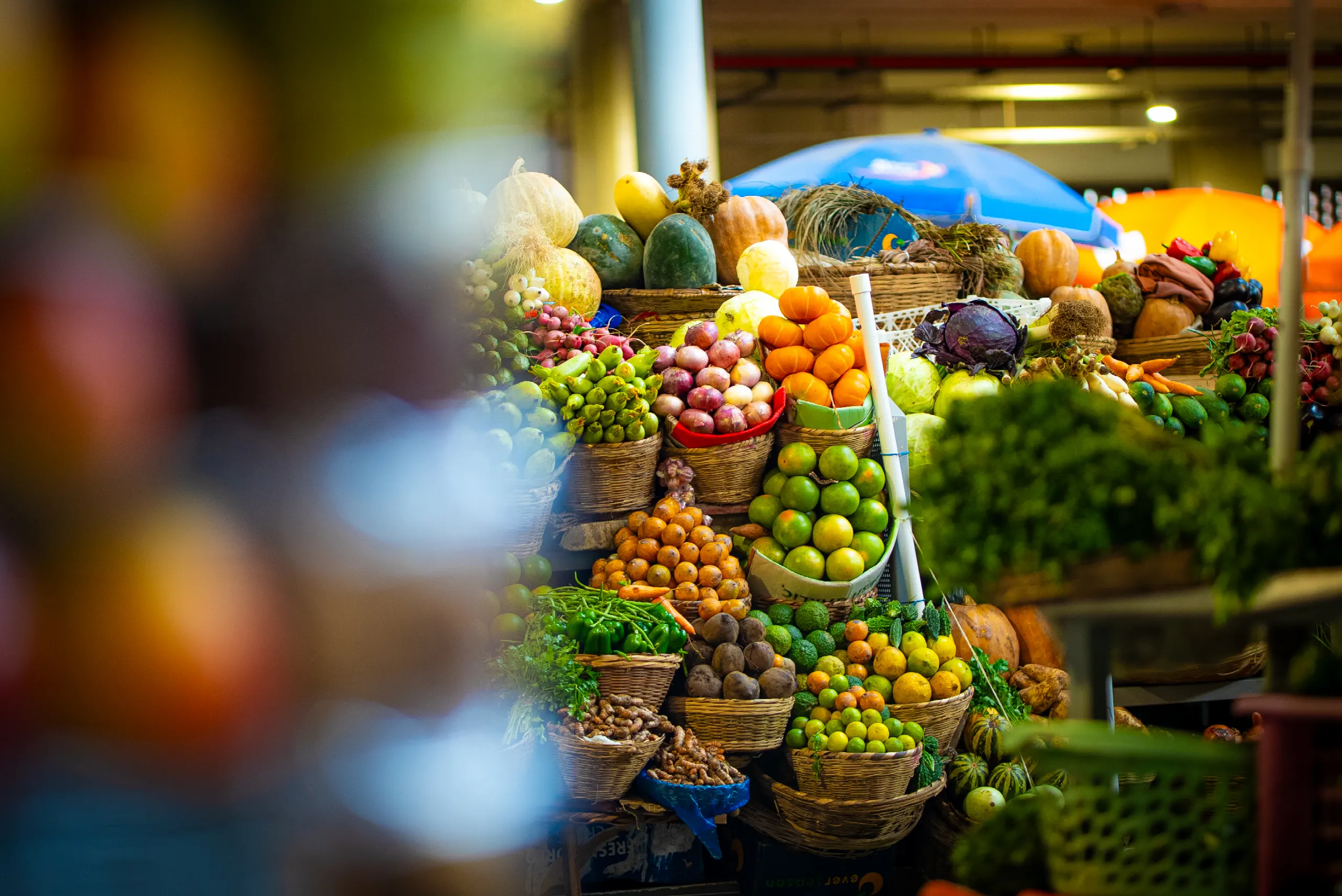 Étals d’un marché couvert garnis de fruits, de légumes et d’herbes fraîches entassés dans des paniers, sous la lumière chaude du hall