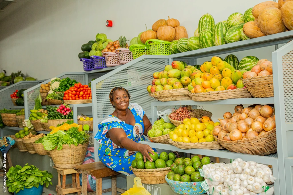 Une vendeuse souriante, en robe à motifs bleus et jaunes, assise entre des étagères de fruits, de légumes, d’oignons et d’ail, une main posée sur des fruits verts