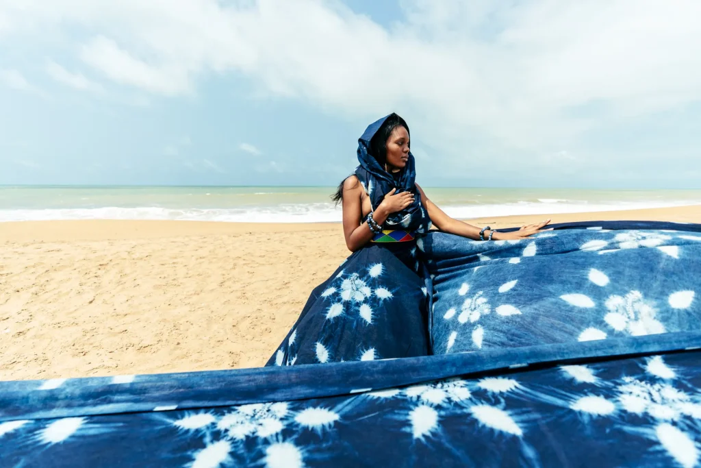 Une femme au foulard bleu, assise sur le sable face à la mer, derrière une étoffe indigo semée de motifs blancs en forme d’étoiles