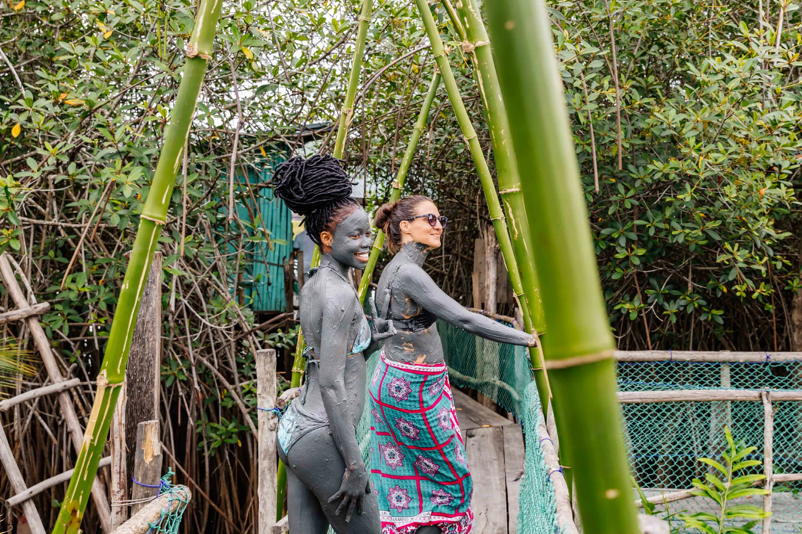 Deux femmes au corps enduit d’argile grise avancent sur une passerelle de bois et de bambou, au milieu de la mangrove