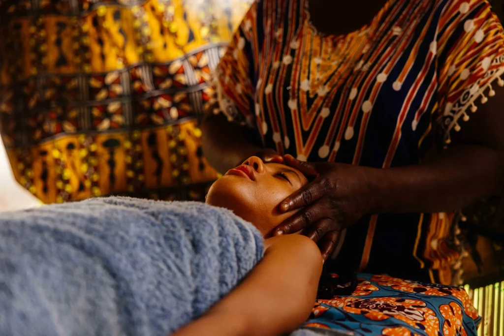 Une masseuse en pagne coloré masse le visage d’une cliente allongée, une serviette bleue sur l’épaule