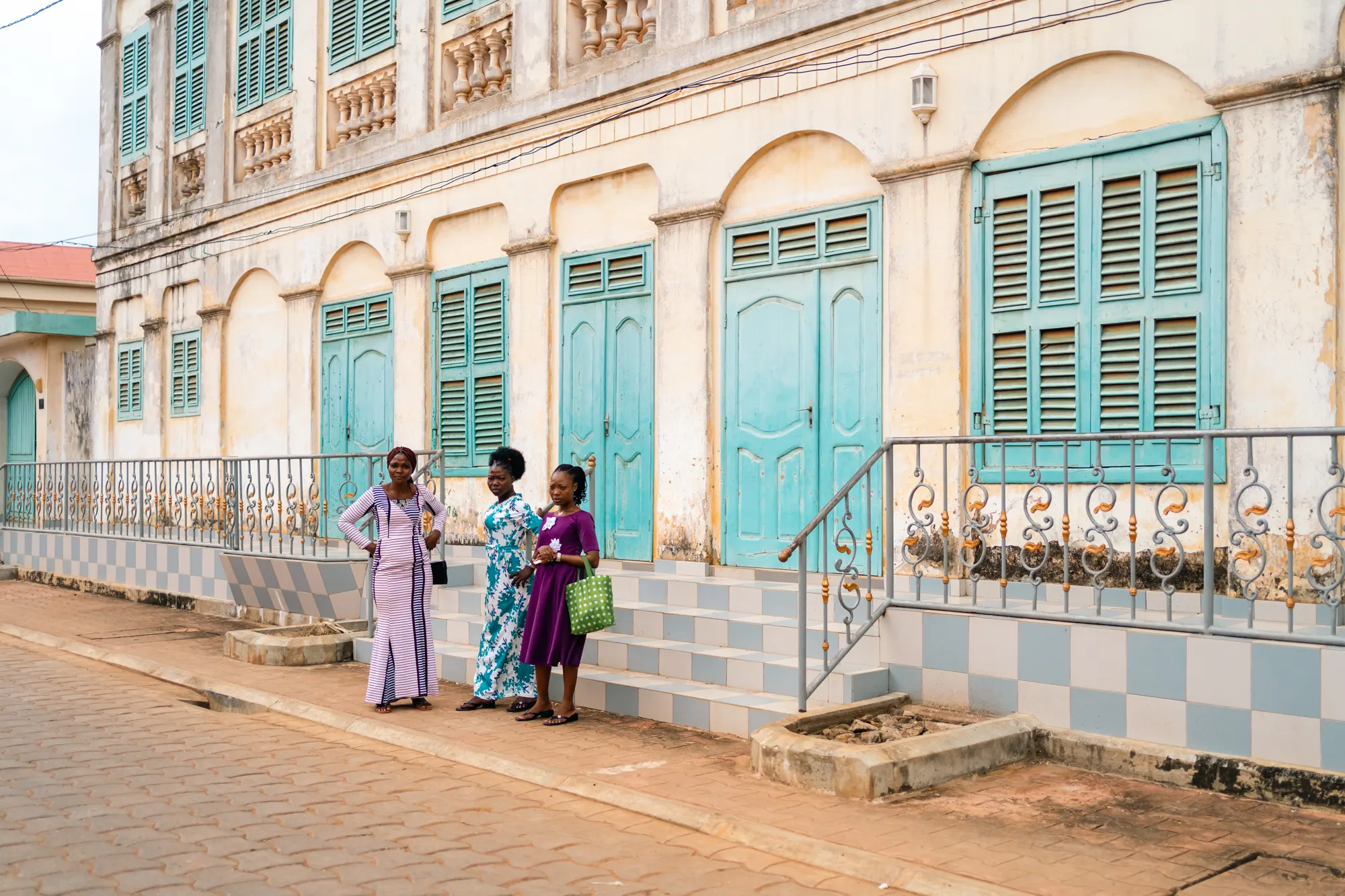 Façade ancienne aux persiennes turquoise et à la ferronnerie ouvragée, devant laquelle trois femmes en tenues colorées discutent sur le trottoir