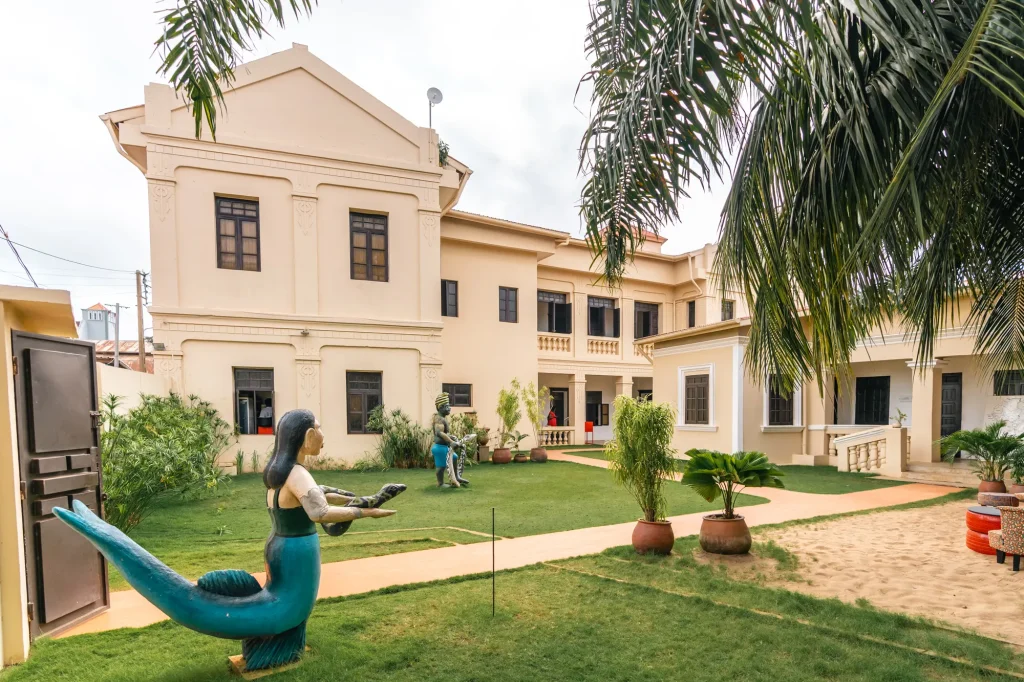 Grande maison crème à étage donnant sur un jardin, avec au premier plan une statue de sirène bleu-vert