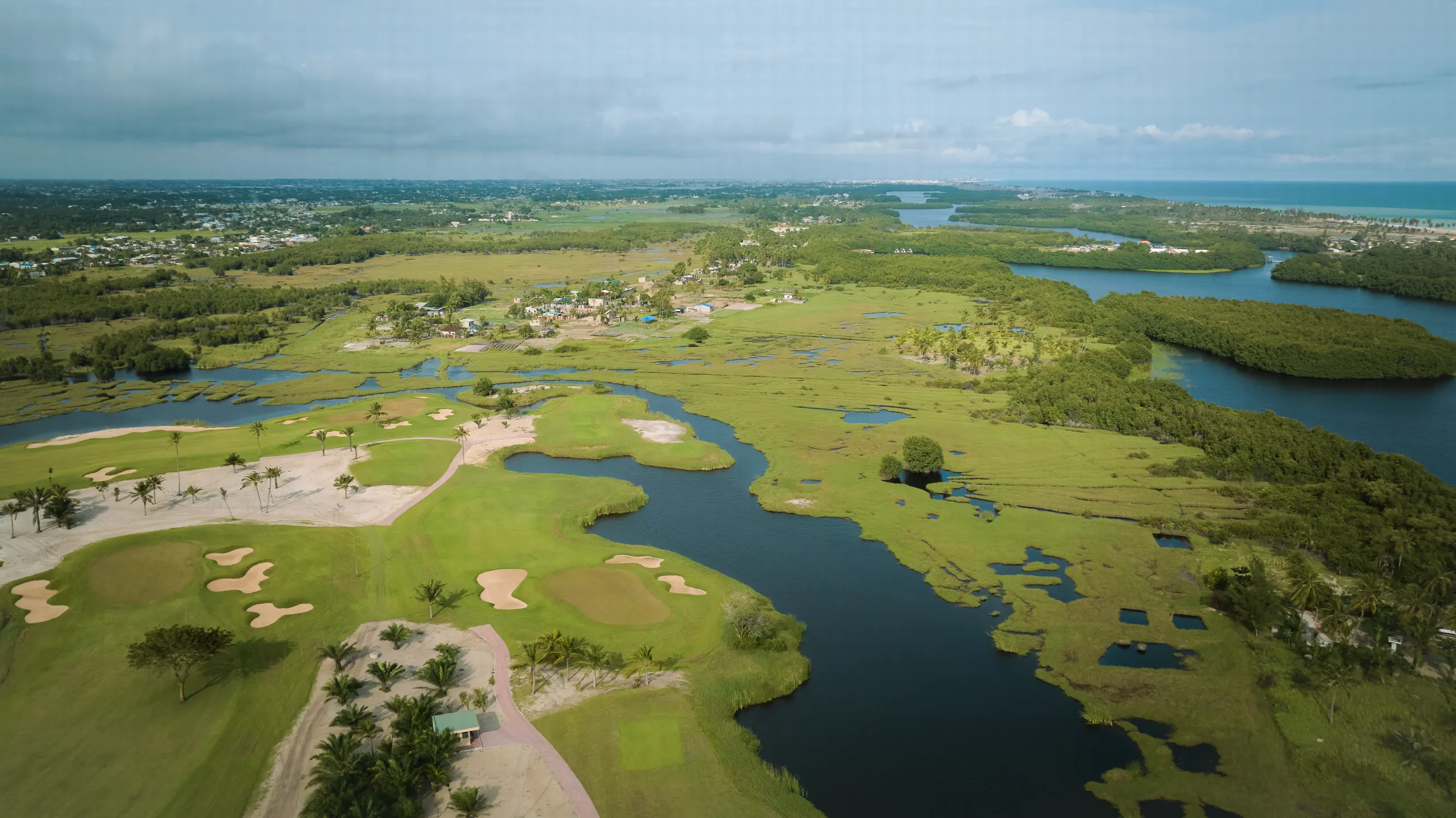 Vue aérienne d’un parcours de golf entre greens, bunkers de sable et plans d’eau, avec la lagune et l’océan à l’horizon