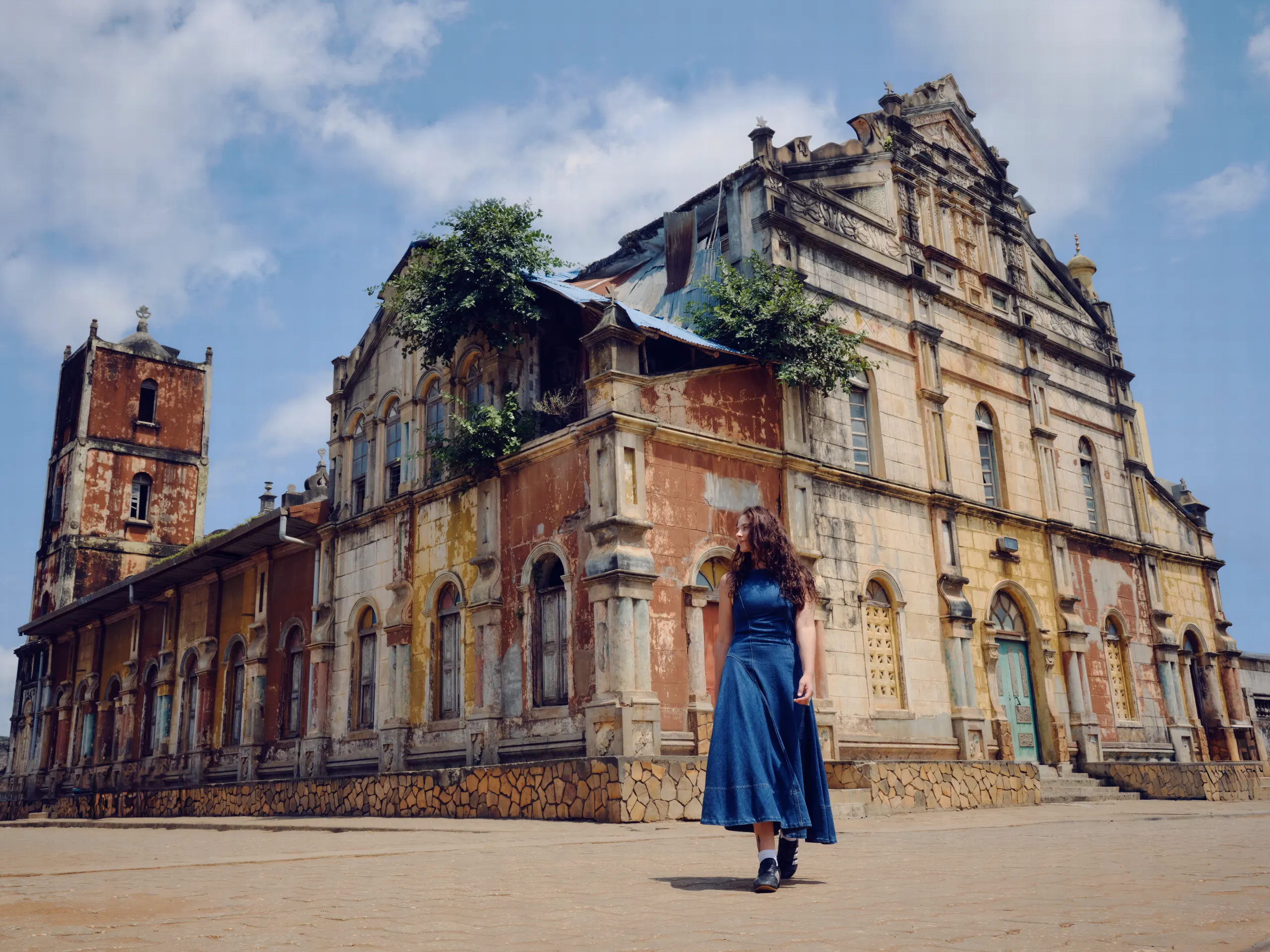 Une femme en robe bleue marche sur la place devant la vaste façade ocre et jaune, patinée par le temps, de la Grande Mosquée, flanquée d’une tour