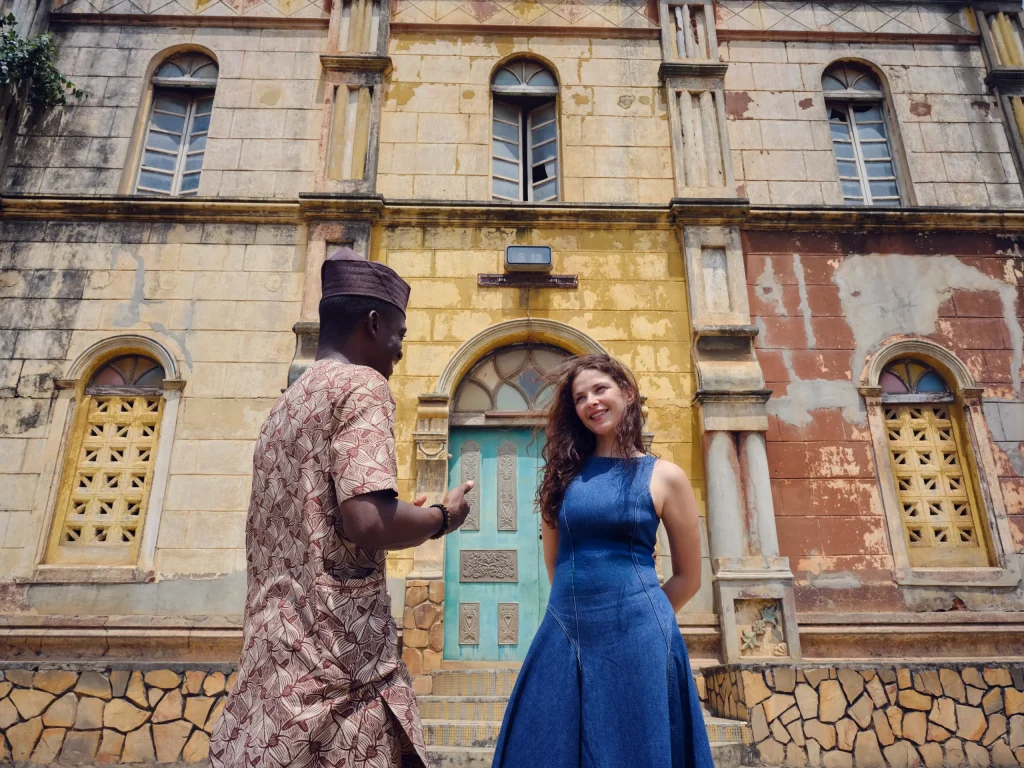 Un guide en tenue traditionnelle s’adresse à une visiteuse devant une façade aux murs jaunes et ocre, à la porte turquoise et aux fenêtres en arc