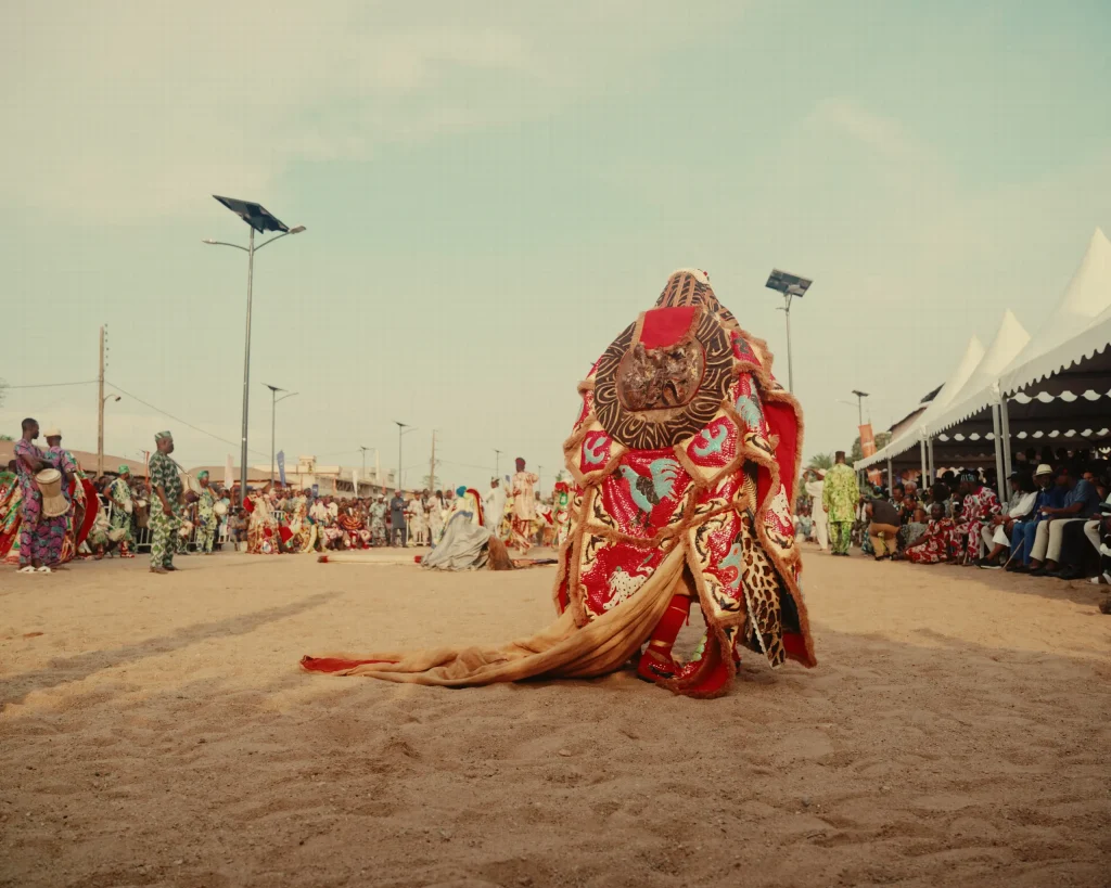 Un masque aux tissus rouges et dorés qui traînent sur le sable, devant des participants rassemblés sous des tentes blanches